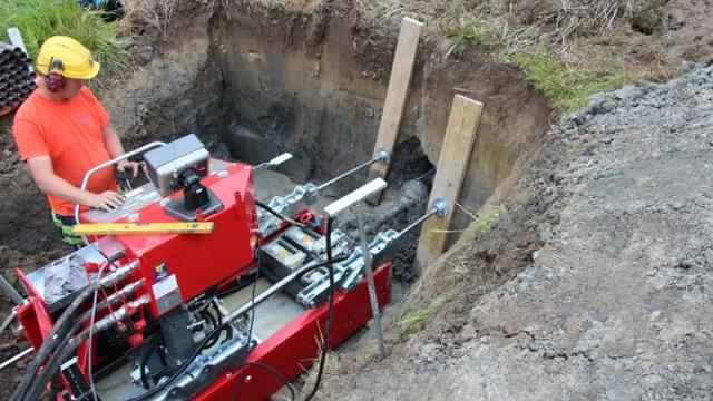 En arbeidstaker i oransje t-skjorte bruker en rød maskin i en grøft for graving og rørlegging.