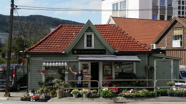 En koselig blomsterbutikk med grønn fasade og rød takstein, omgitt av blomster og planter.