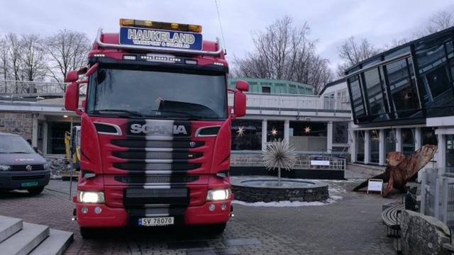 Rød Scania lastebil parkert foran en bygning med moderne arkitektur og en fontene.