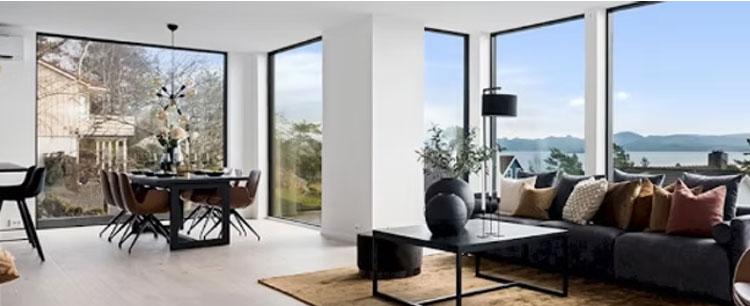 Moderne stue med store vinduer, sofa, spisebord og utsikt over landskapet og vannet.