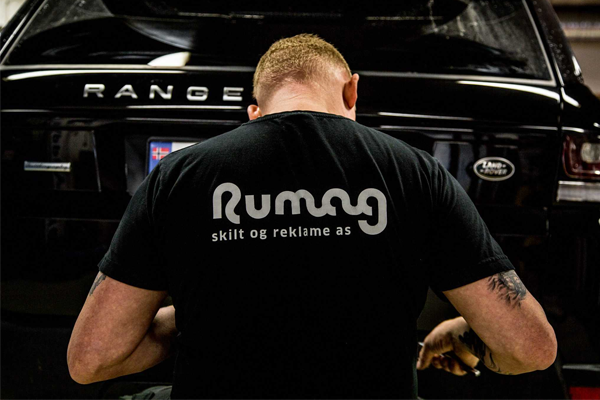 Mann med ryggen til iført svart t-skjorte med teksten "Rumag" foran en svart Range Rover.