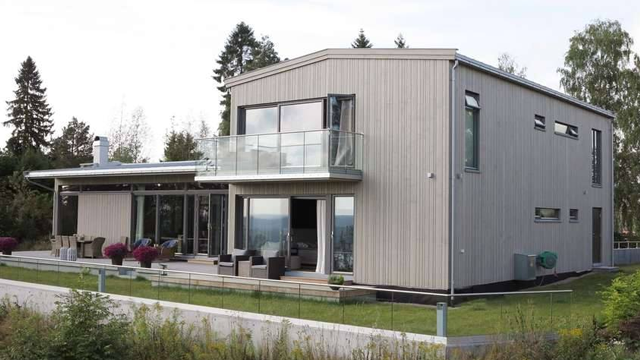 Moderne enebolig med stor terrasse, glassrekkverk og grønne omgivelser.