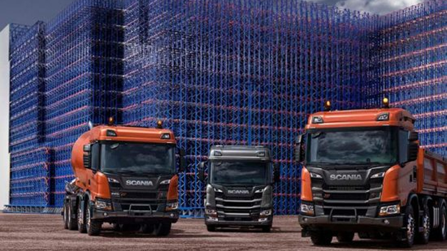 Tre Scania lastebiler står foran en stor bygning med blå og røde stillaser.