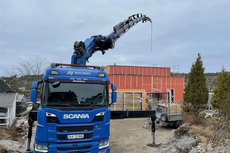 Blå Scania lastebil med kran som løfter materialer ved en byggeplass med rød bygning i bakgrunnen.