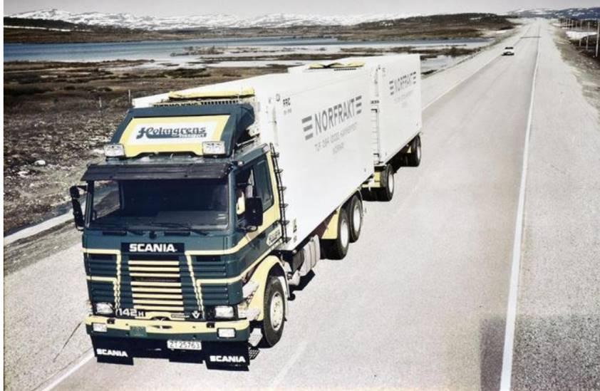 En Scania lastebil med to tilhengere kjører på en vei i et fjellandskap med innsjøer i bakgrunnen.