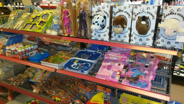 Leketøy og hobbyartikler på en hylle i en butikk, inkludert dukker, spill og kreative sett.