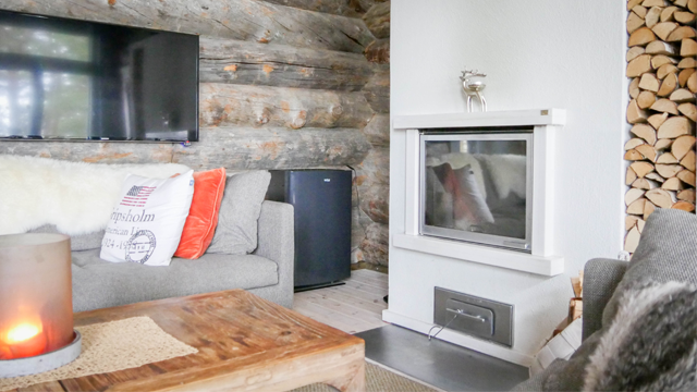 Koselig stue med peis, grå sofa, TV på veggen og vedstabel i bakgrunnen.