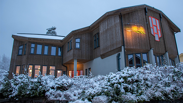 Moderne hus med trepanel og snødekte busker, belyst av en varm glød i skumringen.
