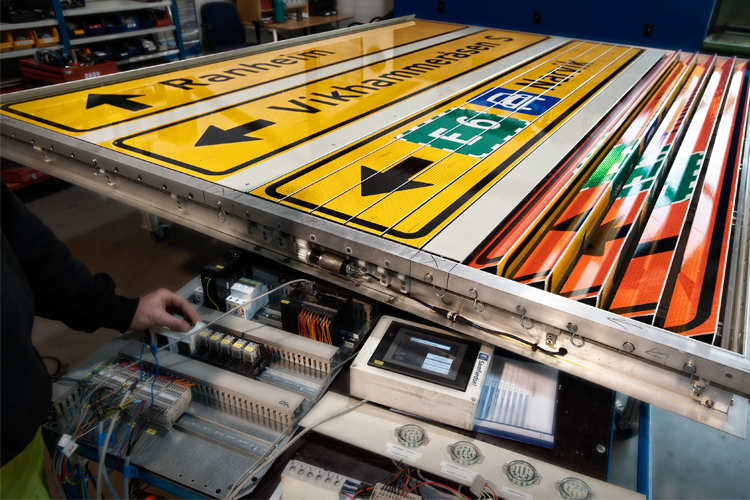 En person justerer elektroniske veiskilt med ulike retninger og informasjon i et verksted.