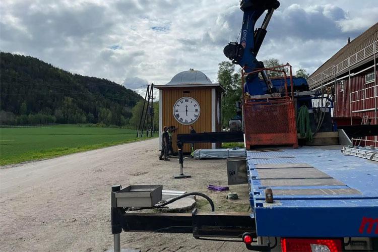 En lastebil med kran står på en grusvei ved en klokke med stor urskive og en bygning i bakgrunnen.
