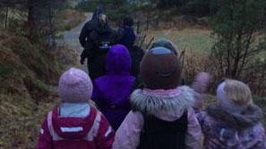 Barn som går på tur i skogen, iført fargerike klær og luer, med en voksen foran dem.