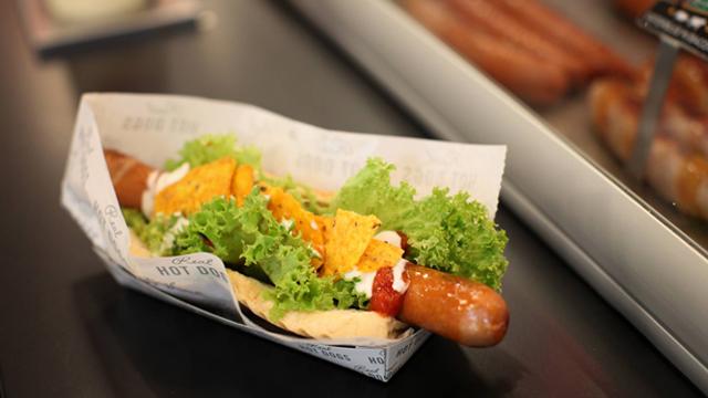 En pølse med salat, ost og dressing i et brød, servert i en papirbeholder.