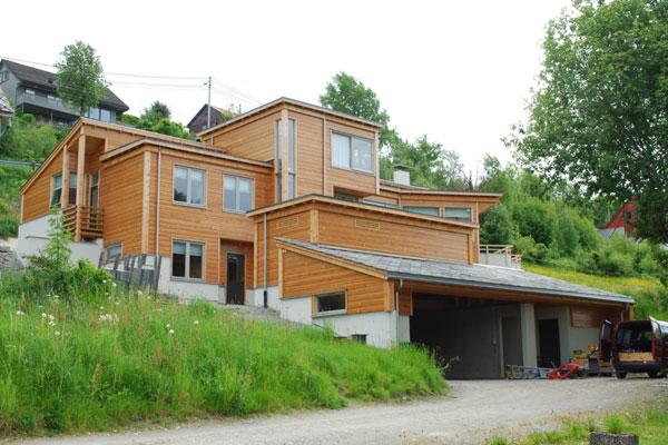 Moderne trehus med flere etasjer, beliggende på en grønn bakke med trær og en grusvei foran.
