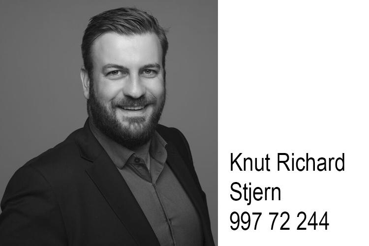 En smilende mann med skjegg iført dress, står mot en grå bakgrunn. Navn og telefonnummer er synlig ved siden av.