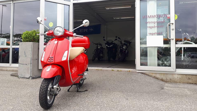 Rød scooter parkert foran en motorsykkelbutikk med flere motorsykler synlige i bakgrunnen.