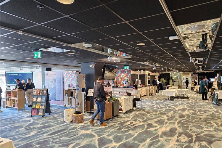 En bokmesse med flere utstillere og besøkende, bøker og stoler i et moderne lokale.