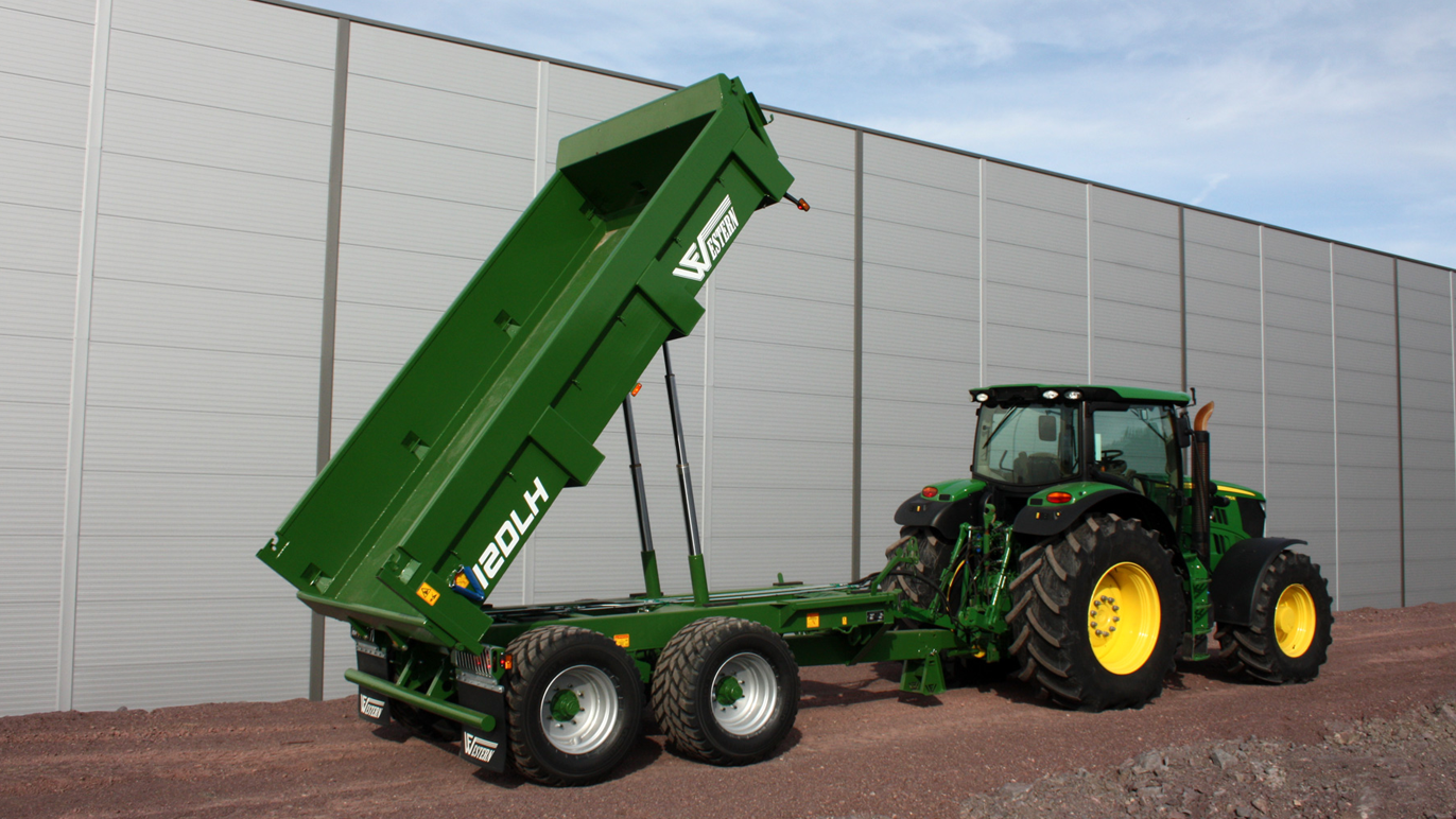Grønn tippvogn koblet til en traktor, hevet for å losse materiale, med en grå bakgrunn.