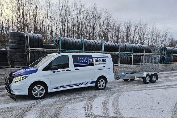 En varebil med tilhenger står parkert ved en dekkstabel på en snødekt vei.