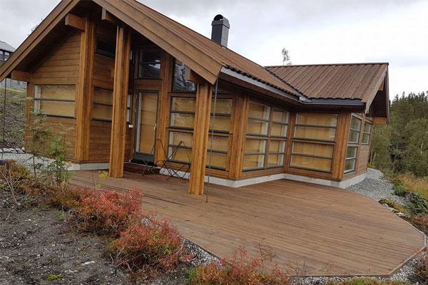 Moderne hytte med trepanel og store vinduer, beliggende i naturskjønne omgivelser. Terrasse i forgrunnen.