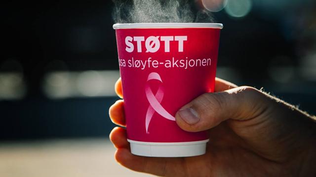 En hånd holder en rosa kopp med teksten "STØTT" og et rosa sløyfe-symbol, damp stiger opp fra koppen.