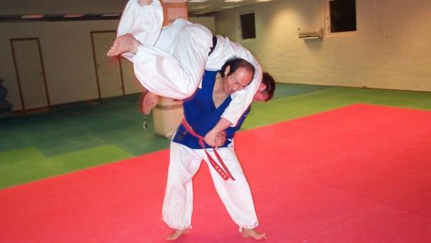 To personer som trener judo, en løfter den andre i en kasteteknikk på en rød og grønn matte.