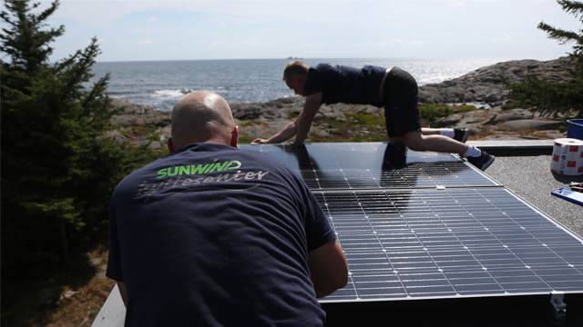 To personer installerer solcellepaneler på et tak med havet i bakgrunnen.