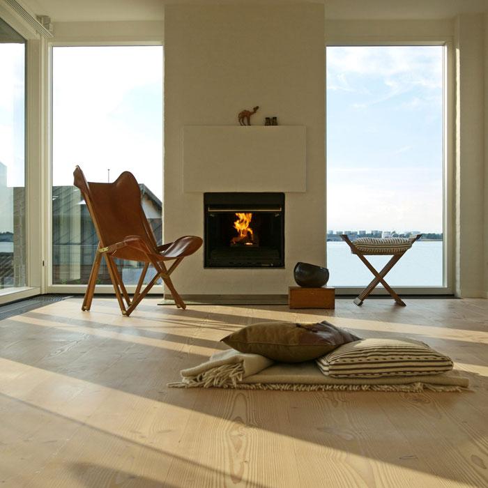 Lyst og moderne stue med peis, lenestol, bord og utsikt over vannet. Sollys strømmer inn gjennom vinduene.