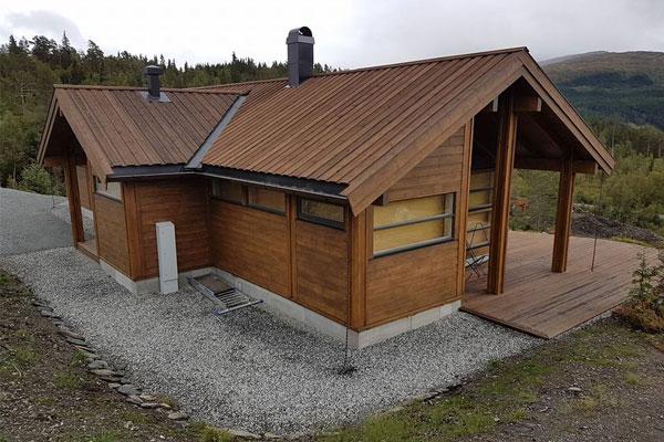 Moderne hytte med trepanel og stor terrasse, omringet av natur i fjellandskap.