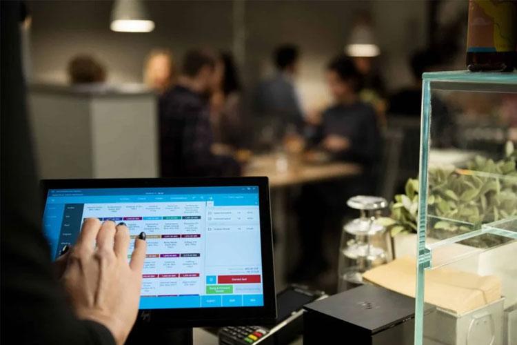 En person bruker en datamaskin med berøringsskjerm i en restaurant, med folk som sitter i bakgrunnen.