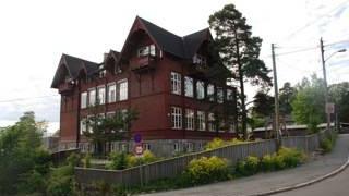 Rødt trehus med skråtak og store vinduer, omgitt av trær og blomster, ligger ved en vei.