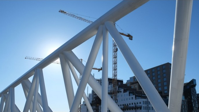 Bilde av en moderne bro med hvite bjelker, med byggekraner og en klar blå himmel i bakgrunnen.