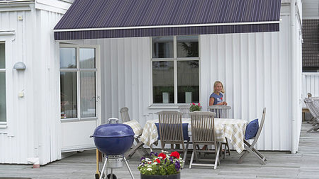 En kvinne står ved et bord på en terrasse med en grill og blomsterpotter i solfylt vær.