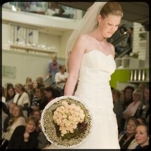 Brud med blomsterbukett går nedover catwalken, omgitt av publikum som ser på.