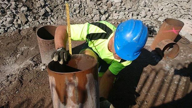 En arbeidstaker i sikkerhetsutstyr måler en rusten rørdel på en byggeplass med stein i bakgrunnen.