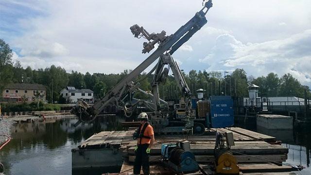 En arbeider står på en båt ved en havn, med en kran og bygninger i bakgrunnen under en skyet himmel.
