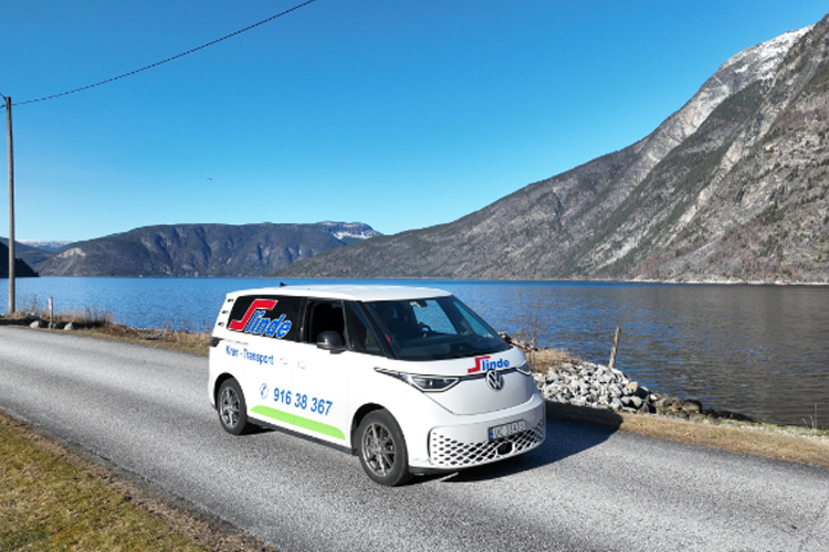 En varebil med logoen til et transportselskap kjører langs en vei ved en innsjø med fjell i bakgrunnen.