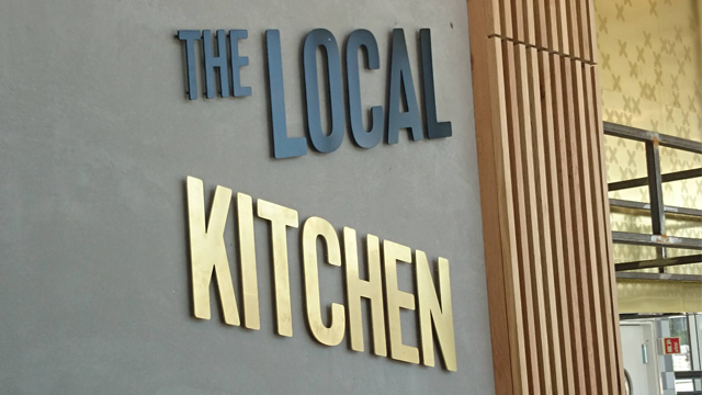 Skilt med teksten "The Local Kitchen" i moderne design på en vegg.