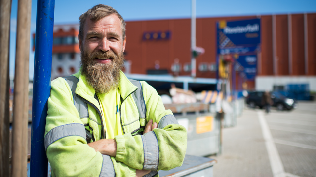 En smilende mann med skjegg iført refleksjakke står foran en byggeplass med containere og en butikk i bakgrunnen.