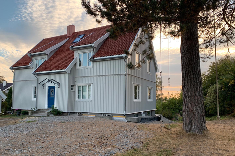 Moderne enebolig med rød takstein og grå panel, omgitt av trær og en solfylt himmel.