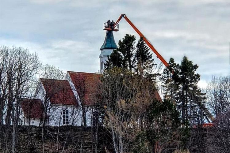 Kirke med heisekran som arbeider på taket, omringet av trær og en grå himmel.