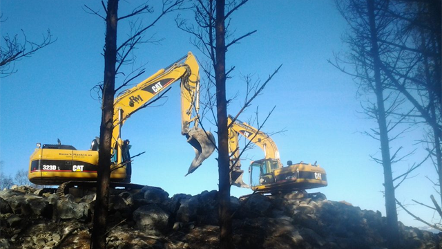 To store og gule gravemaskiner arbeider på en steinete bakke under en klar blå himmel.