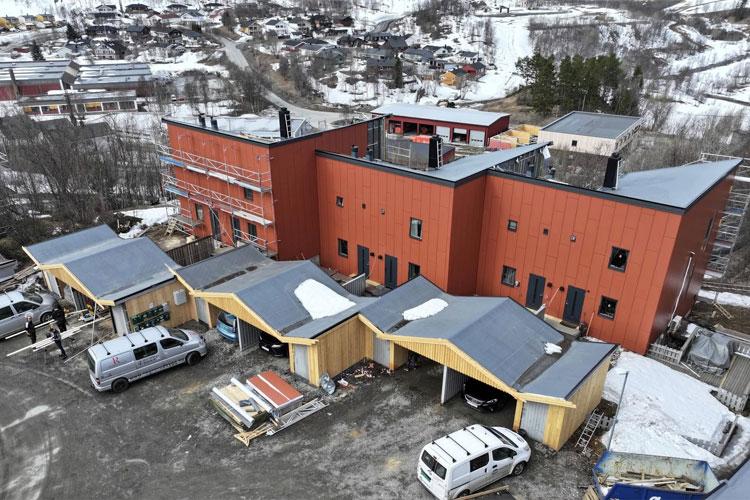 Moderne bygning med rød fasade og tak i snøfylt landskap, med parkerte biler og byggearbeid i nærheten.