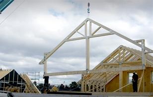 Bygging av hus med trestendere, kran som løfter takstoler, og arbeidere på byggeplassen under en skyet himmel.