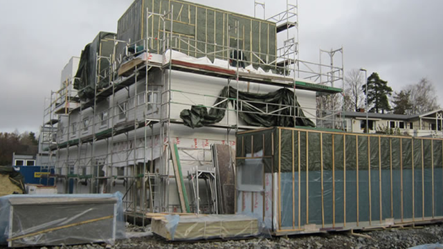 Bygning under oppføring med stillas, plastdeksel og materialer på byggeplassen.