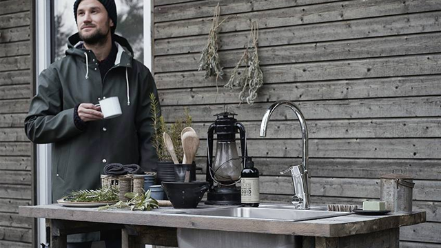 En mann står ved en utendørs kjøkkenbenk med en kopp i hånden, omgitt av urter og kjøkkenutstyr.