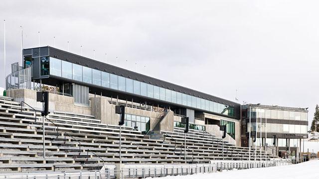 Moderne bygning med glassfasade og tribuner, plassert i et åpent område med grå himmel.