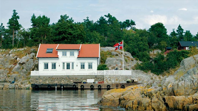 Hvit hytte med rød tak på en klippe ved sjøen, med norsk flagg og trær i bakgrunnen.