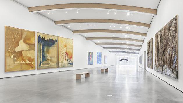 Moderne kunstgalleri med store malerier på veggene og en benk i midten av rommet. Lys og åpen atmosfære.