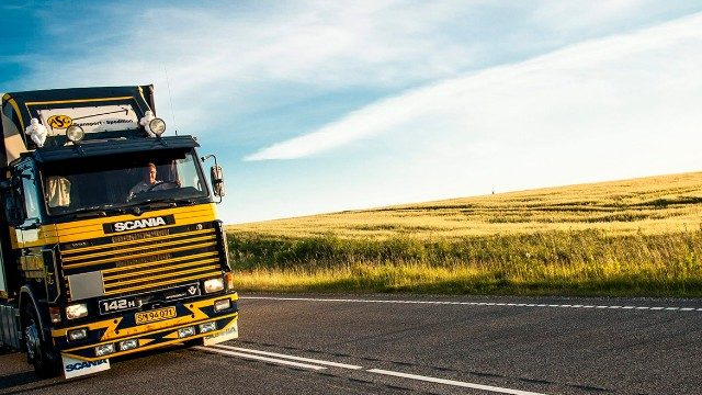 En stor Scania lastebil kjører på en landevei med grønne åser i bakgrunnen under en blå himmel.