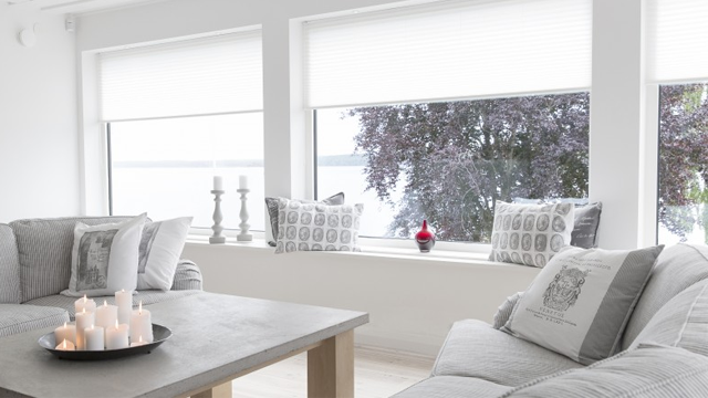 Lyst stue med grå sofaer, et bord med stearinlys og vinduer som viser utsikt mot vannet og trær utenfor.